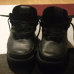 Svarta sneakers med snörning - Säljer ett par helsvarta sneakers med klassisk design och rund tå. Skorna har snörning och är tillverkade i syntetmaterial med perforerade detaljer på ovandelen. Perfekta för en stilren och enkel look.