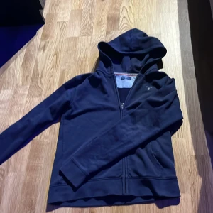 Mörkblå hoodie från GANT - Säljer en stilren mörkblå hoodie från GANT med dragkedja och broderad logga på bröstet. Tröjan har huva och långa ärmar, perfekt för en avslappnad look. Klassisk design med ribbade muddar och mjukt material.