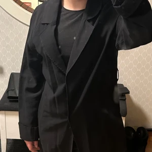 Svart lång kappa från Vero Moda - Säljer en svart lång kappa från Vero Moda. Klassisk design med krage och långa ärmar. Perfekt för dig som gillar stilrena och tidlösa plagg. Passar bra till både jeans, kjol och klänning. Midjeband finns ej! Använd fåtal gånger
