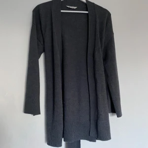 Mörkgrå kofta i merinoull med knytband - Säljer en mörkgrå lång kofta från Cubus i 100% merinoull. Koftan har långa ärmar, öppen front och ett knytband i midjan som ger en snygg siluett. Perfekt för lager-på-lager och riktigt mjuk och skön.