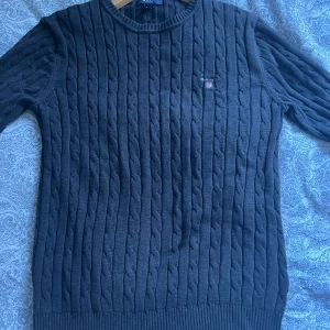 Mörkgrå kabelstickad tröja från GANT - Säljer en mörkgrå kabelstickad tröja från GANT med rund halsringning och diskret logga på bröstet. Den har används runt 10 gånger men har ett riktigt fint skick. Storleken är L men passade perfekt till mig som har M