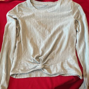 Turkos ribbad långärmad blus från Lindex - Säljer en ljusgrå ribbad långärmad topp från Lindex med knutdetalj framtill. Perfekt för dig som gillar enkla men stilrena plagg. Mjuk och stretchig, passar dig som har storlek 158/164.