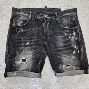 Svarta jeansshorts från Dsquared2 M/48 - Säljer ett par snygga svarta jeansshorts från Dsquared2 med slitna detaljer, hål och färgstänk. Shortsen har fem fickor, knappgylf och uppvikta benslut. Perfekta för en avslappnad stil.