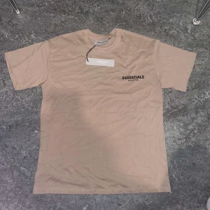 Beige t-shirt från Essentials Fear of God - Snygg beige t-shirt från Essentials Fear of God med svart logga tryckt på bröstet och etikett i nacken. Klassisk rund hals och normal passform. Perfekt för en stilren och avslappnad look.