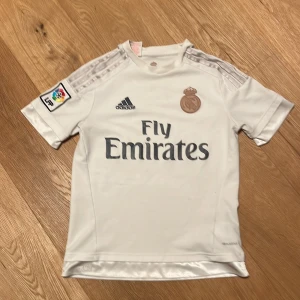 Vit Real Madrid fotbollströja från Adidas - Säljer en vit Real Madrid fotbollströja från Adidas med Fly Emirates-tryck på bröstet och klubbmärke i guld. Tröjan har korta ärmar och La Liga-märke på ena ärmen. Perfekt för fotbollsfans!