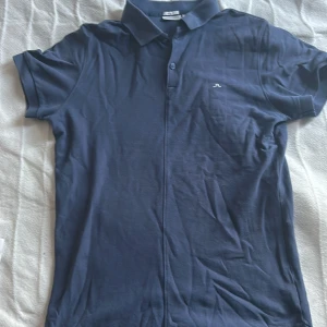 Blå piké från J.Lindeberg - Säljer en stilren blå pikétröja från J.Lindeberg i slim fit-modell. Tröjan har korta ärmar, klassisk krage och knappar framtill. Liten broderad logga på bröstet. Perfekt för en clean och sportig look. Perfekt nu när golfsäsongen börjar!