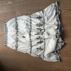 Vit långkjol med broderade detaljer - Säljer en vit långkjol med svarta och bruna broderade blommor och dekorativa band. Kjolen har volanger och knytband med pärlor i midjan. Perfekt för dig som gillar bohemisk stil och vill ha något unikt i garderoben.