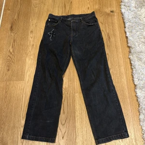 Svarta jeansbyxor med tryck från Lukamachain - Unika svarta jeansbyxor från Lukamachain med stora vita tryck på baksidan där det står 'CHAIN WEAR'. Klassisk femficksmodell med dragkedja och raka ben. Perfekta för dig som vill sticka ut med din stil.