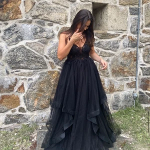 Svart långklänning med spets från Mascara London - Säljer nu min balklänning från Mascara London med v-ringad spetsliv och volangig kjol i flera lager tyll. Klänningen har öppen rygg och vackra spetsdetaljer upptill, perfekt för bal eller fest. Enbart använd en gång (bal). Storlek 40 (skulle säga ”M”). Nypris 3045kr.