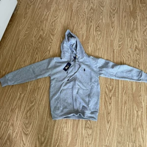 Grå hoodie med dragkedja från Polo Ralph Lauren - Säljer en klassisk grå hoodie från Polo Ralph Lauren med dragkedja framtill och huva. Tröjan har två fickor och den ikoniska lilla broderade loggan på bröstet. Perfekt för en avslappnad och stilren look.