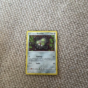 Pokémonkort Galarian Stunfisk - Säljer ett Pokémonkort med Galarian Stunfisk. Kortet har en gul ram och motivet visar Galarian Stunfisk omgiven av gröna detaljer. Kortet har 100 HP och attacker som Field Trap och Tackle. Perfekt för samlare eller Pokémonfans!