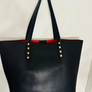 Svart shoppingväska med nitar från Even&Odd - Stilren svart shoppingväska från Even&Odd med guldfärgade nitar på handtagen och röd insida. Väskan har en innerficka med dragkedja och tryckknappsstängning upptill. Perfekt för dig som vill ha plats med allt du behöver.