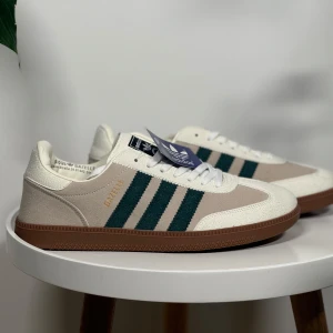 Adidas Gazelle sneakers med gröna ränder - Snygga Adidas Gazelle sneakers i beige mocka med klassiska gröna ränder på sidorna och vit ovandel. Skorna har brun gummisula och vit snörning. Perfekta för dig som gillar retrostil och vill ha något stilrent till vardags. Finns i storlek 41 & 45