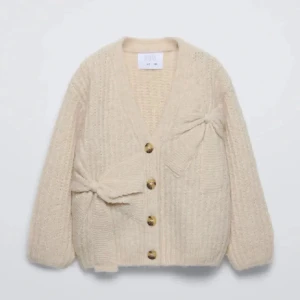 Beige kofta med rosetter från Zara - Superfin beige stickad kofta från Zara med två stora rosetter framtill. Koftan har v-ringning, knappar och ribbad struktur. Perfekt att styla till jeans eller kjol för en trendig look.