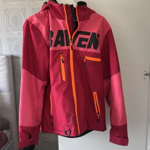 Röd skaljacka från Raven Snowmobile Gear - Skaljacka från Raven. Jackan har huva, flera fickor med dragkedja och är perfekt för utomhusaktiviteter. Storlek M