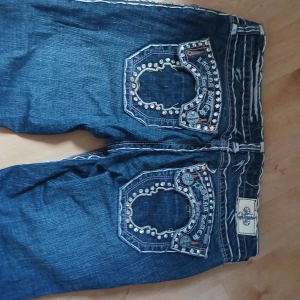 Blå jeans med dekorerade bakfickor - Säljer ett par blå jeans med unika, dekorerade bakfickor med nitar och broderier. Jeansen har normal passform och klassisk femficksdesign. Perfekta för dig som vill sticka ut med detaljer på dina byxor.