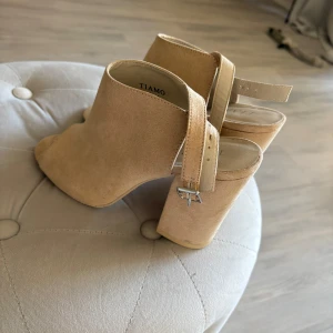 Beiga klackskor från Tiamo - Säljer ett par snygga beiga klackskor från Tiamo med öppen tå och bred klack. Skorna har en justerbar rem med spänne runt hälen och är tillverkade i mockaliknande material. Några fläckar men utöver de fint i skick 