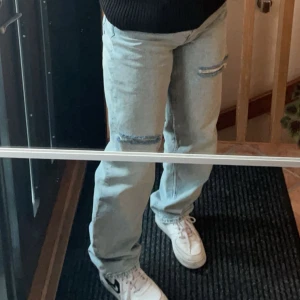 Ljusblå slitna jeans med knappar - Säljer ett par ljusblå jeans med slitningar på knäna och knäppning med flera silverfärgade knappar. Jeansen har en rak passform .