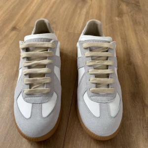 Maison margiela gats helt nya - Snygga sneakers i vitt och ljusgrått med ovandel i skinn och mocka. Skorna har beige snörning och en klassisk gummisula. Perfekta för en stilren och avslappnad look.