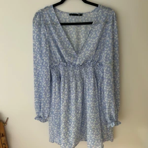 Ljusblå blommig klänning från 157 - Supersöt ljusblå klänning med vitt blommönster från 157. Klänningen har v-ringning, smock i midjan och långa ärmar med volangdetalj vid ärmslut. Perfekt för vår och sommar!