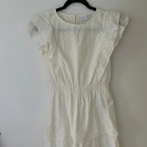 Vit kortklänning med broderade blommor från VILA - Söt vit klänning från VILA i storlek 36 med broderade små blommor, volangdetaljer vid axlarna och resår i midjan. Klänningen har kort ärm och är tillverkad i ett lätt material, perfekt för varma dagar.