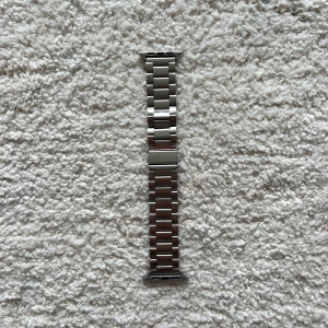 Silver länkarmband till Apple Watch - Säljer ett stilrent silverfärgat länkarmband i rostfritt stål till Apple Watch. Armbandet har en klassisk design med breda länkar och en diskret låsdel där det står 'Apple Watch'. Perfekt för dig som vill ge din klocka en exklusiv look.