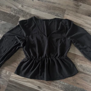 Svart blus med v-ringning från Vero Moda - Säljer en svart blus från Vero Moda med v-ringning och långa ärmar. Blusen har markerad midja med resår och knappar vid ärmsluten. Aldrig använd