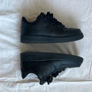 Svarta Nike Air Force 1 sneakers - Säljer ett par helsvarta Nike Air Force 1 sneakers med klassisk design. Skorna har snörning, rund tå och platt sula. Perfekta för dig som gillar stilrena och tidlösa sneakers. Nike-loggan syns på sidan och tungan.