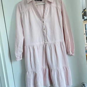 Ljusrosa klänning från Zara - Supersöt ljusrosa kort klänning från Zara med volanger och knappar framtill. Klänningen har lång ärm och en avslappnad passform, perfekt för vår och sommar. Tillverkad i ett mjukt bomullsmaterial.