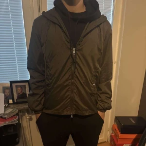 Mörkgrön Moncler windbreaker  - Säljer en grön vindjacka från Moncler med huva och dragkedja framtill. Jackan har Moncler-logga på ärmen och en rödvit rand på axeln. Perfekt för dig som vill ha en stilren och sportig look. Säljer den på grund av att den inte passar mig längre. Hör av dig om du är intresserad!