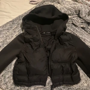 Svart croppad dunväst med huva från Zara - Säljer en svart croppad dunväst från Zara med huva och dragkedja. Västen har vadderad front och rygg samt släta ärmar i sweatshirtmaterial. Perfekt för lager-på-lager och streetstyle.