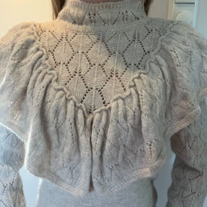 Beige stickad tröja med volang från Dilvin - Superfin beige stickad tröja från Dilvin med höghals, volangdetaljer och vackert hålmönster. Tröjan har långa ärmar och en feminin, romantisk look. Perfekt att styla till jeans eller kjol för en trendig outfit. Köpte för 499kr