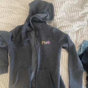 Mörkgrå hoodie från PINK - Mysig mörkgrå hoodie från PINK med dragkedja framtill och huva med dragsko. Broderad logga i färgglada bokstäver på bröstet. Perfekt för en avslappnad stil.