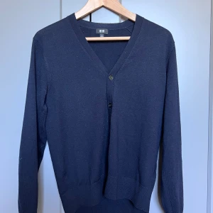 Mörkblå kofta från Uniqlo - Säljer en klassisk mörkblå kofta från Uniqlo med v-ringning och knäppning framtill. Tröjan är långärmad och tillverkad i ett mjukt, tunt material som passar perfekt till lager-på-lager. Enkel och stilren design.