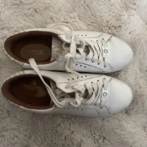 Vita sneakers från Michael Kors - Vita sneakers från Michael Kors med beige logotypdetaljer baktill och på plösen. Skorna har snörning och rund tå, samt diskreta perforeringar på sidorna. 