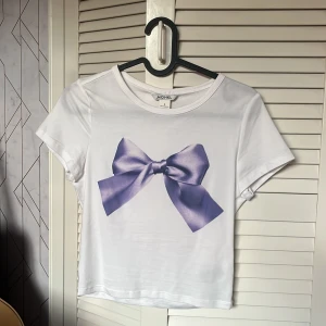 Vit t-shirt med lila rosett från Monki - Vit t-shirt från Monki med ett stort tryck av en lila rosett på framsidan. T-shirten har korta ärmar och en klassisk rund halsringning. Perfekt för dig som vill ha en söt och trendig look. Använd en gång, inga tecken på användning, nypris var 149kr.