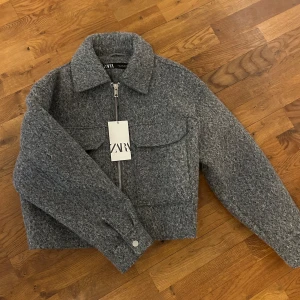 Grå ulljacka från Zara - Säljer en grå, croppad jacka från Zara i ullblandning. Jackan har dragkedja framtill, stora bröstfickor och ballonärmar. Aldrig använd!