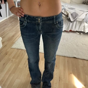 Mörkblå lowwaist bootcut jeans - Säljer ett par mörkblå jeans med låg midja och bootcut passform. Jeansen har snygga broderade detaljer på bakfickorna och dubbla knappar framtill. Klassisk denim med lätt slitning och dekorativa sömmar😚