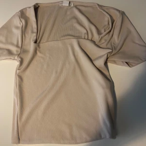 Beige ribbad topp med fyrkantig ringning - Säljer en stilren beige topp med korta ärmar och fyrkantig ringning. Toppen är ribbad och har en enkel, clean look som passar perfekt till jeans eller kjol. Materialet känns mjukt .