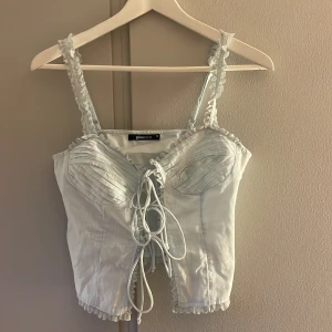 Ljusblå korsettopp från Gina Tricot - Supersöt ljusblå korsettopp från Gina Tricot med spetsdetaljer och volang längs axelbanden. Toppen har snörning framtill och veckade kupor för extra stil. 