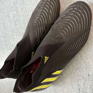 Adidas Predator Edge+ FG fotbollsskor - Säljer ett par svarta Adidas Predator Edge+ FG fotbollsskor med gula detaljer och röda dobbar. Skorna har en strumpliknande passform utan snörning och är tillverkade i syntetmaterial. Perfekta för dig som vill ha grym kontroll och stil på planen. Fotbollsskor har använts 2 gånger 
