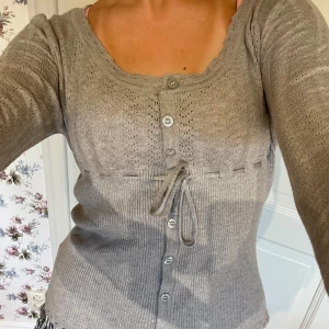 Grå kofta med knyt och knappar - Supersöt grå kofta med lång ärm, knappar hela vägen och knytband i midjan. Fint hålmönster upptill och ribbad nedtill, samt söt kant runt hals och nederkant. Perfekt för lager-på-lager och chill vibes.