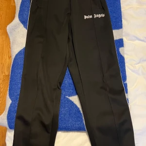 Svarta byxor från Palm Angels - Svarta trackpants från Palm Angels med vit logga tryckt på ena sidan och vita ränder längs benen. Byxorna har en rak passform och är tillverkade i ett glansigt syntetmaterial som ger en sportig vibe.