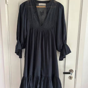 Mörkblå klänning från Lolly's Laundry - Supersnygg mörkblå klänning från Lolly's Laundry med v-ringning, broderade detaljer och volang nertill. Klänningen har trekvartsärmar med volang och en loose passform. Perfekt för dig som gillar en avslappnad men chic stil.