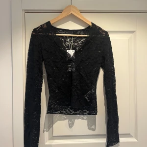 Svart spetsblus från Gina Tricot - Supersnygg svart långärmad blus från Gina Tricot i genombruten spets med v-ringning. Toppens spetsmönster ger en cool och elegant vibe, perfekt att styla med jeans eller kjol. Figurnära passform och transparent känsla för en trendig look.