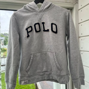 Grå hoodie från Polo Ralph Lauren - Snygg grå hoodie från Polo Ralph Lauren med broderad POLO-text i svart och vitt framtill. Klassisk känguruficka, huva och ribbade muddar. Perfekt för chill dagar och enkel att matcha med jeans eller joggers. Den har ett litet streck vitt färg på magen, sista bilden. Storlek S typ