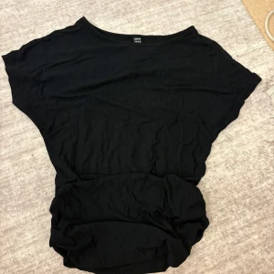 Svart loose topp med rynkade detaljer - En svart loose-fit topp med bred halsringning och rynkade detaljer i midjan. Den är ganska lång men men som sagt så har den rynkade sidor.