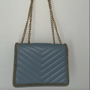 Ljusblå quiltad axelväska med kedja - Snygg blå axelväska med quiltat mönster och guldfärgad kedja som axelrem. Väskan har en stilren, rektangulär form och detaljer i metall runt kanterna. Perfekt för dig som vill ha en trendig och praktisk väska med coola detaljer.