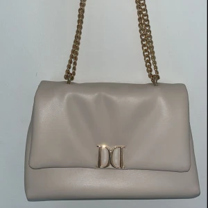  superfin beige axelväska med guldfärgad kedja!  - En stilren och lyxig väska från Don Donna som passar till både vardags och fest. Mjuk design i beige konstläder med ett snyggt gulddetalj-spänne framtill (dubbel-D). Kedjan gör att den känns riktigt lyxig. Dubbelfack och ett fack med dragkedja invändigt. Använd endast en gång 