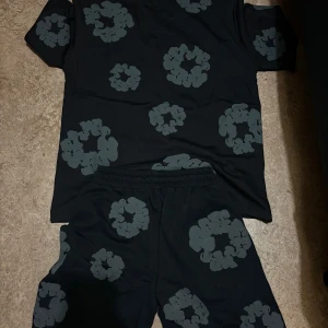 Denim tears tracksuit/ set - Svart t-shirt från Denim Tears med gråa blommiga tryck över hela plagget. Klassisk passform med rund hals och korta ärmar. Tillverkad i mjuk bomull, perfekt för en chill och trendig look.